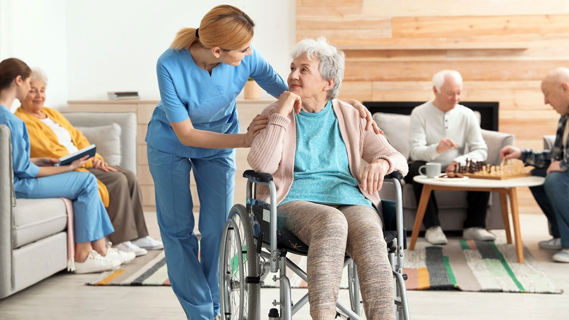 Healthcare worker talking with senior patient in wheelchair, her hands on the patient's shoulders. In the background is another worker talking with an elderly female and two elderly men playing chess.